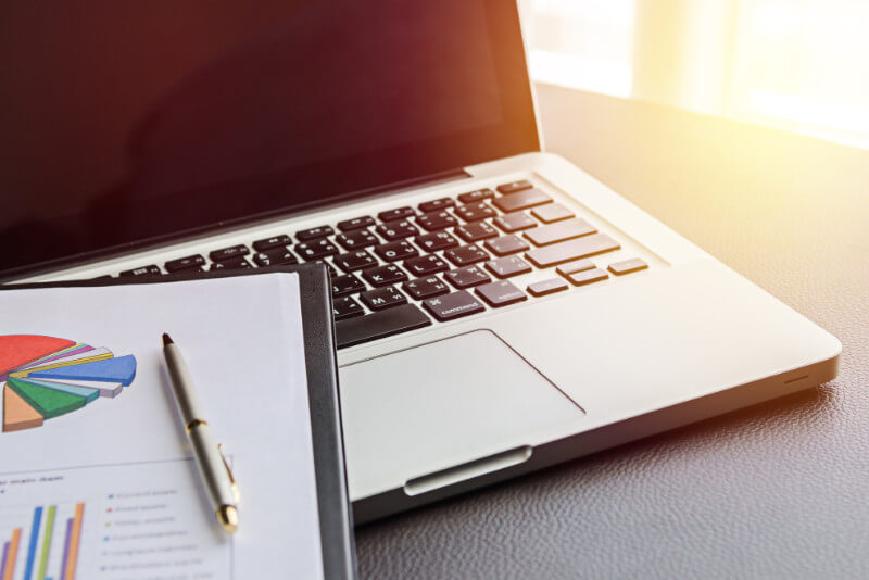 A laptop on a desk with some financial reports on top of it.