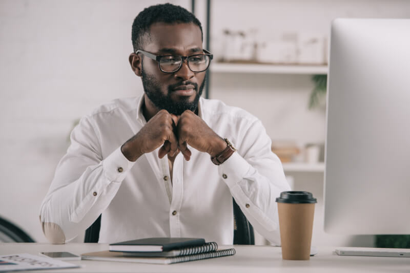 An African American man sitting at a computer in an office with a cup of coffee and a notebook.